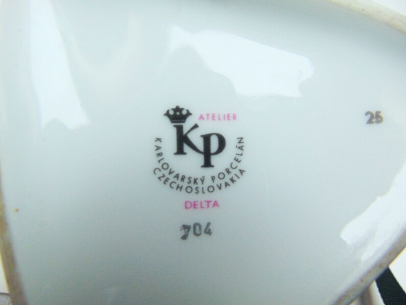 Może przedstawiać: Biały talerz porcelanowy z czarno-r&oacute;żowym logo, na kt&oacute;rym widnieje napis "Atelier KP Karlovarsky Porcelain Czechoslovakia Delta 704" oraz numer "25" w prawym g&oacute;rnym rogu.