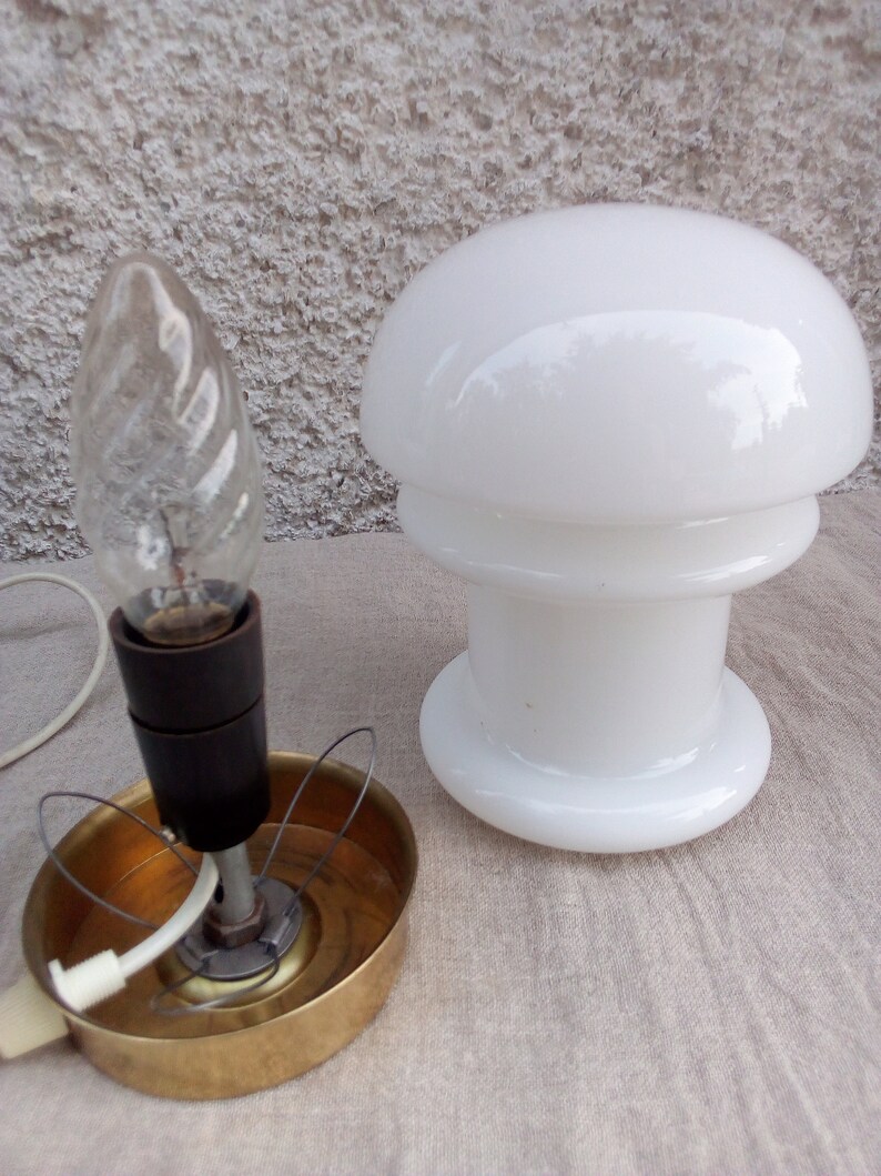 May include: A white glass mushroom-shaped lampshade with a brass base and a white cord. The lampshade has a double rim and a smooth, rounded top. The base has a wire cage around the bulb.