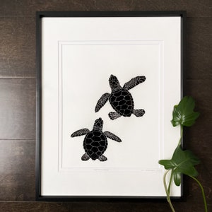 Puede incluir: L&aacute;mina art&iacute;stica enmarcada con dos cr&iacute;as de tortuga marina negras sobre fondo blanco. Las tortugas est&aacute;n ilustradas con patrones detallados en el caparaz&oacute;n. La impresi&oacute;n est&aacute; en un marco negro con un passe-partout blanco. El texto "Hatchlings" es visible.