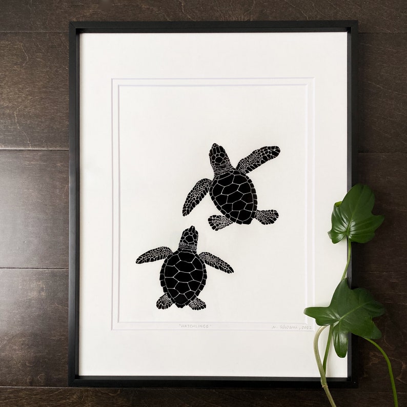 Puede incluir: L&aacute;mina art&iacute;stica enmarcada con dos cr&iacute;as de tortuga marina negras sobre fondo blanco. Las tortugas est&aacute;n ilustradas con patrones detallados en el caparaz&oacute;n. La impresi&oacute;n est&aacute; en un marco negro con un passe-partout blanco. El texto "Hatchlings" es visible.