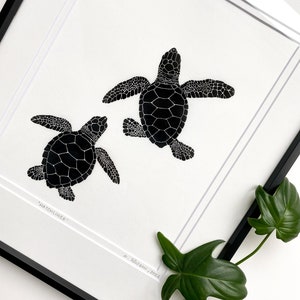 Puede incluir: L&aacute;mina art&iacute;stica enmarcada en blanco y negro con una ilustraci&oacute;n de dos tortugas marinas. Las tortugas est&aacute;n representadas en una ilustraci&oacute;n detallada y estilizada, con la palabra "Hatchlings" impresa en la parte inferior. La obra est&aacute; firmada y fechada.