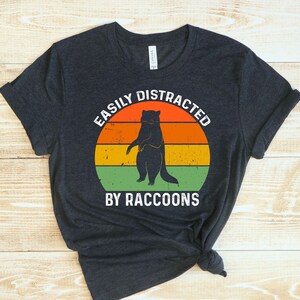 Op de afbeelding: Een donkergrijs T-shirt met een vintage-stijl afbeelding van een wasbeer. De afbeelding zegt "Easily Distracted by Raccoons".