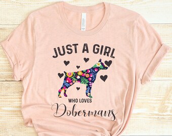 May include: A light pink t-shirt with the text "JUST A GIRL WHO LOVES Dobermans". The shirt features a floral patterned Doberman silhouette with black hearts.