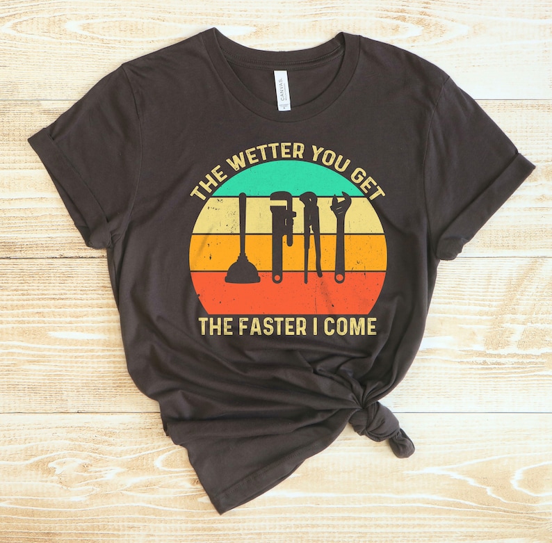 Puede incluir: Camiseta gris oscuro con un dise&ntilde;o retro de puesta de sol y herramientas de fontaner&iacute;a. El texto dice: "THE WETTER YOU GET THE FASTER I COME".