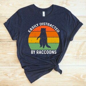 Op de afbeelding: Een marineblauw T-shirt met een vintage-stijl grafische afbeelding van een wasbeer die op zijn achterpoten staat. De afbeelding is omgeven door een cirkel met de tekst "Easily Distracted by Raccoons".