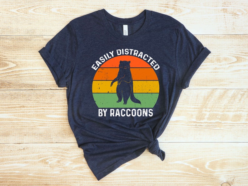 Op de afbeelding: Een marineblauw T-shirt met een vintage-stijl grafische afbeelding van een wasbeer die op zijn achterpoten staat. De afbeelding is omgeven door een cirkel met de tekst "Easily Distracted by Raccoons".