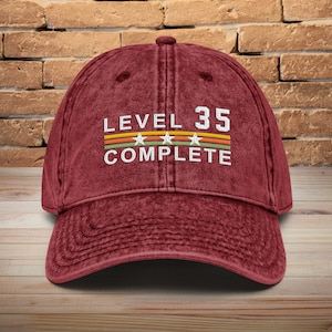 May include: A maroon baseball cap with the text "LEVEL 35 COMPLETE" in white with a colorful stripe design and three white stars.