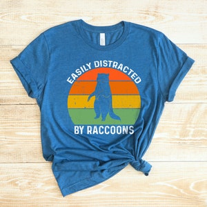 Op de afbeelding: Een blauw T-shirt met een vintage-stijl afbeelding van een wasbeer die voor een zonsondergang staat. De tekst "Easily Distracted By Raccoons" is op het shirt gedrukt.
