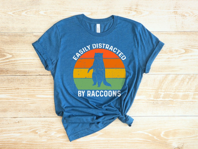 Op de afbeelding: Een blauw T-shirt met een vintage-stijl afbeelding van een wasbeer die voor een zonsondergang staat. De tekst "Easily Distracted By Raccoons" is op het shirt gedrukt.