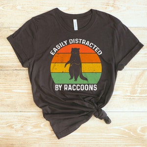 Op de afbeelding: Een donkerbruin T-shirt met een vintage-stijl grafische afbeelding van een wasbeer die op een zonsondergangachtergrond staat. De tekst "Easily Distracted By Raccoons" is in wit onder de afbeelding gedrukt.