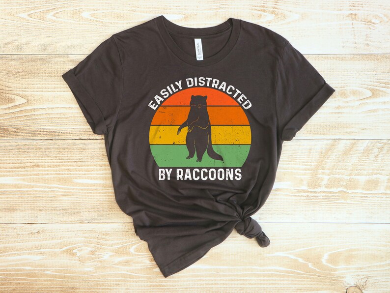 Op de afbeelding: Een donkerbruin T-shirt met een vintage-stijl grafische afbeelding van een wasbeer die op een zonsondergangachtergrond staat. De tekst "Easily Distracted By Raccoons" is in wit onder de afbeelding gedrukt.