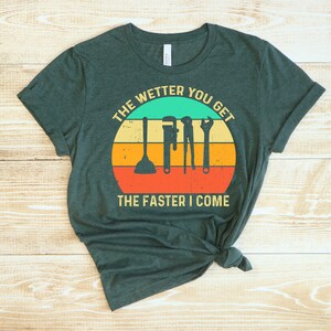 Puede incluir: Camiseta verde oscuro con un gr&aacute;fico que presenta herramientas de fontaner&iacute;a dentro de un dise&ntilde;o de puesta de sol. El texto dice "THE WETTER YOU GET THE FASTER I COME". La camiseta es de material suave y tiene cuello redondo.