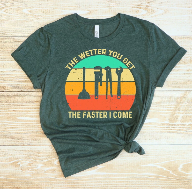 Puede incluir: Camiseta verde oscuro con un gr&aacute;fico que presenta herramientas de fontaner&iacute;a dentro de un dise&ntilde;o de puesta de sol. El texto dice "THE WETTER YOU GET THE FASTER I COME". La camiseta es de material suave y tiene cuello redondo.