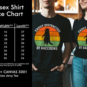Op de afbeelding: Unisex zwart T-shirt met een retro-grafiek van een wasbeer en de tekst "Easily Distracted by Raccoons". Maattabel inbegrepen met metingen in inches.