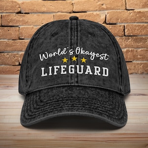 May include: Black vintage washed cotton baseball cap with white embroidered text "World's Okayest Lifeguard" and three yellow stars.