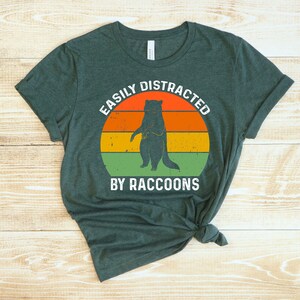 Op de afbeelding: Een donkergroen T-shirt met een vintage-stijl grafische afbeelding met de tekst "Easily Distracted by Raccoons". De afbeelding toont een wasbeer die op een zonsondergangachtergrond staat met oranje, gele en groene strepen.