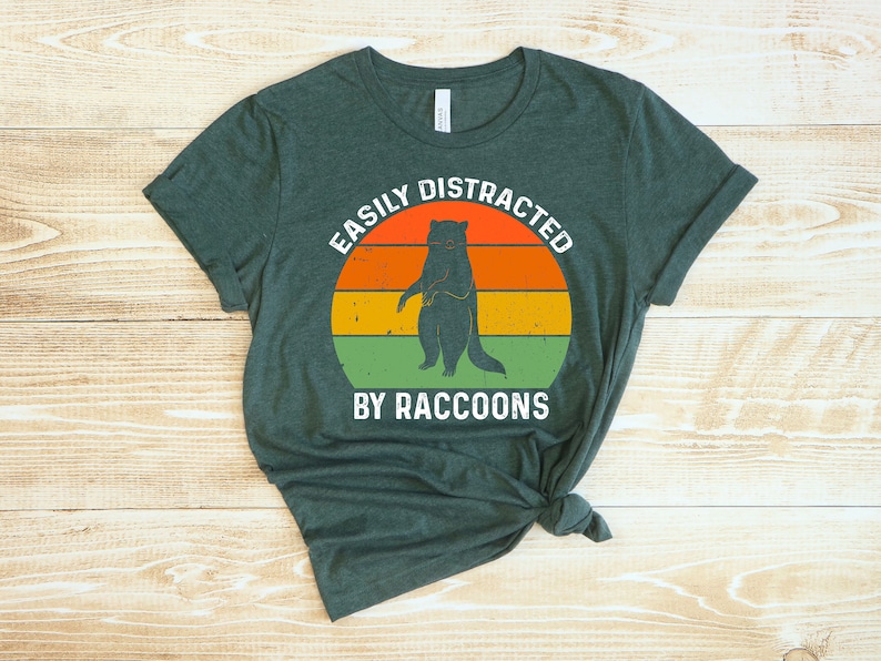Op de afbeelding: Een donkergroen T-shirt met een vintage-stijl grafische afbeelding met de tekst "Easily Distracted by Raccoons". De afbeelding toont een wasbeer die op een zonsondergangachtergrond staat met oranje, gele en groene strepen.