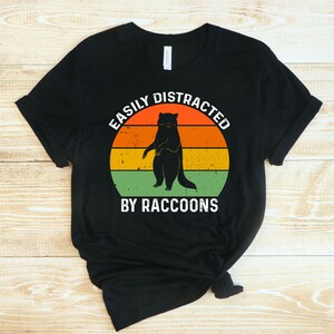 Op de afbeelding: Zwart T-shirt met een retro-stijl grafische afbeelding van een wasbeer die voor een zonsondergang staat. De tekst "Easily Distracted By Raccoons" is onder de afbeelding gedrukt.