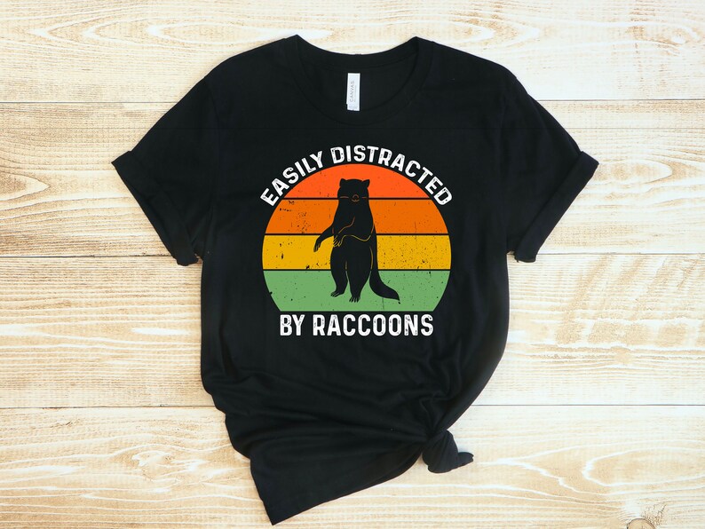 Op de afbeelding: Zwart T-shirt met een retro-stijl grafische afbeelding van een wasbeer die voor een zonsondergang staat. De tekst "Easily Distracted By Raccoons" is onder de afbeelding gedrukt.