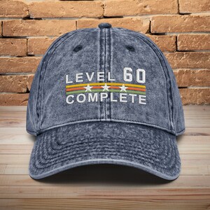May include: A blue denim baseball cap with the text "LEVEL 60 COMPLETE" in white with a colorful stripe design and white stars.