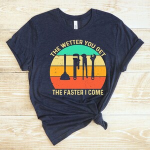 Puede incluir: Camiseta azul marino con un dise&ntilde;o retro de puesta de sol. El dise&ntilde;o presenta herramientas de fontaner&iacute;a y el texto "THE WETTER YOU GET THE FASTER I COME". La camiseta est&aacute; hecha de un material suave y c&oacute;modo.