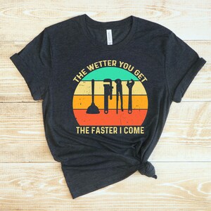 Puede incluir: Camiseta gris oscuro con un gr&aacute;fico circular que presenta herramientas de fontaner&iacute;a y el texto "THE WETTER YOU GET THE FASTER I COME" en un esquema de colores retro. La camiseta est&aacute; anudada en la parte inferior.