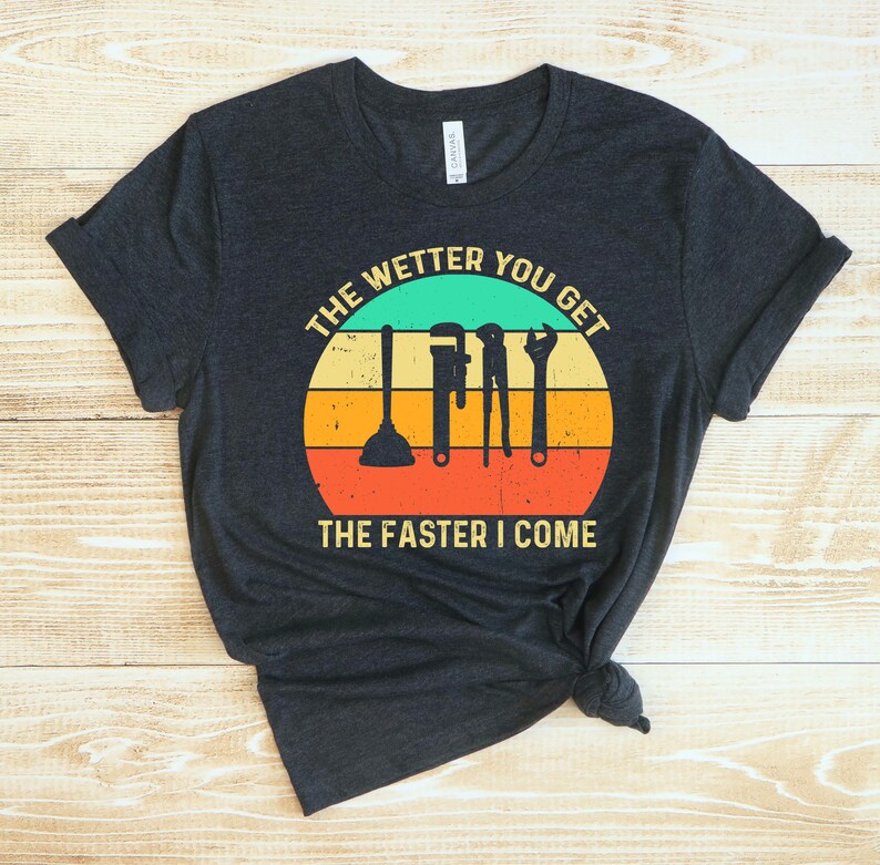 Puede incluir: Camiseta gris oscuro con un gr&aacute;fico circular que presenta herramientas de fontaner&iacute;a y el texto "THE WETTER YOU GET THE FASTER I COME" en un esquema de colores retro. La camiseta est&aacute; anudada en la parte inferior.