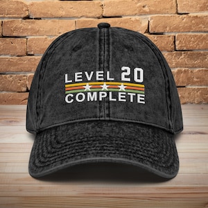 May include: Black vintage style baseball cap with the text "LEVEL 20 COMPLETE" in white with a yellow, orange, green, and white stripe design.