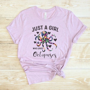 Puede incluir: Una camiseta de color lavanda con el texto "Just a girl who loves Octopuses" en negro. Un dise&ntilde;o de pulpo floral est&aacute; centrado en la camiseta.