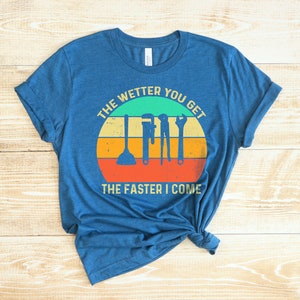 Puede incluir: Camiseta azul con un dise&ntilde;o gr&aacute;fico que presenta herramientas de fontaner&iacute;a y el texto "THE WETTER YOU GET THE FASTER I COME" en un dise&ntilde;o retro de puesta de sol. La camiseta est&aacute; anudada en la parte inferior.