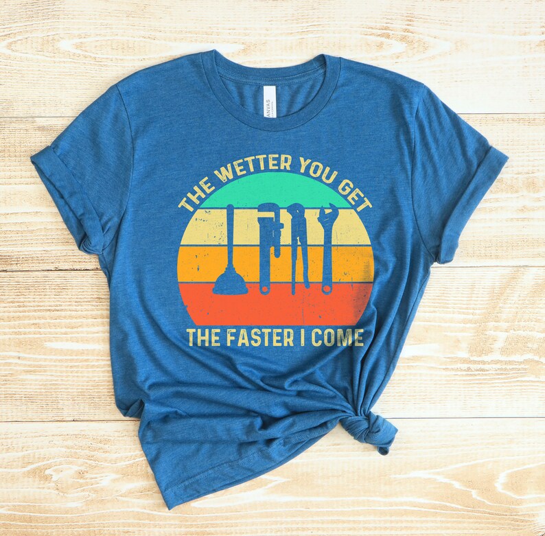 Puede incluir: Camiseta azul con un dise&ntilde;o gr&aacute;fico que presenta herramientas de fontaner&iacute;a y el texto "THE WETTER YOU GET THE FASTER I COME" en un dise&ntilde;o retro de puesta de sol. La camiseta est&aacute; anudada en la parte inferior.