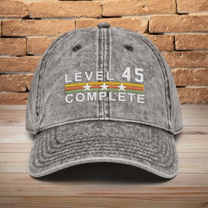 May include: A gray distressed denim baseball cap with the text "LEVEL 45 COMPLETE" in white with three white stars and a multi-colored stripe.