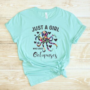 Puede incluir: Una camiseta turquesa con el texto "Just a girl who loves Octopuses" en negro. Un dise&ntilde;o de pulpo floral est&aacute; centrado en la camiseta.