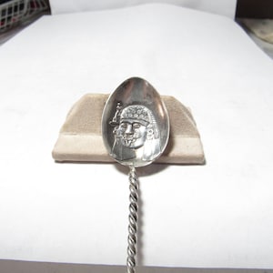 May include: A silver spoon with a detailed face design on the bowl. The handle is twisted. The spoon is resting on a beige surface against a white background. The face appears to be of a historical figure.