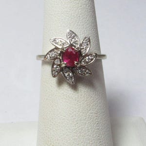 May include: A silver-toned ring featuring a central round, deep pinkish-red gemstone surrounded by a cluster of small, clear, sparkling stones arranged in a petal-like, floral pattern. The band is thin and metallic, supporting the decorative setting.