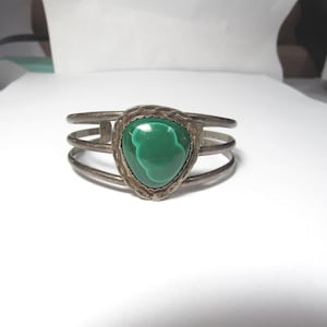 May include: Silver cuff bracelet with a large, teardrop-shaped, vibrant green malachite stone. The bracelet has three parallel bands and a textured bezel setting around the stone. A piece of jewellery.