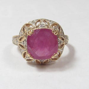 May include: A ring featuring a large, cushion-cut, vibrant pink gemstone set in a gold and silver-toned metal frame. The setting is decorated with intricate filigree patterns and small, clear, sparkling stones along the band and halo.