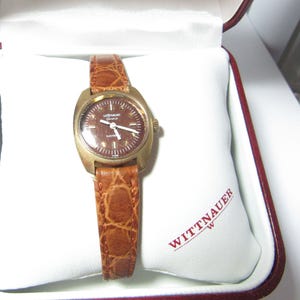 May include: A vintage Wittnauer Genève Electric watch with a brown face and a gold-toned case. The watch has a brown leather band with a crocodile pattern. The watch is displayed in a white box with the Wittnauer logo.