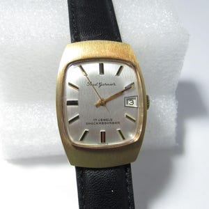 May include: A vintage Paul Garnier wristwatch with a gold-toned rectangular case and a silver dial. The watch features gold hands, a date window, and the text "17 JEWELS SHOCKABSORBER". It has a black leather strap.