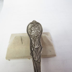 May include: Silverware spoon with a detailed relief design. The handle features a Native American profile, surrounded by corn stalks and leaves. The spoon is likely made of sterling silver.