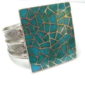 May include: A silver cuff bracelet with a large square turquoise mosaic design. The turquoise stones are set in a silver frame, and the bracelet band has a textured, ridged design.