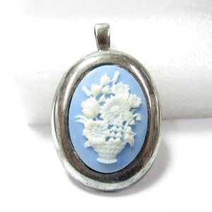May include: Oval pendant with a light blue cameo featuring a white floral basket design. The cameo is set in a silver-toned metal frame with a loop at the top for a chain.