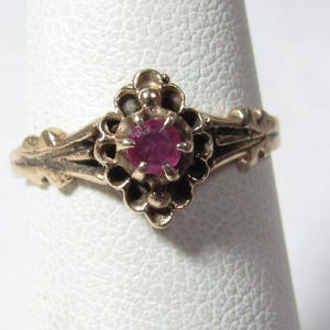 May include: A gold-toned ring featuring a central, round, deep red gemstone. The ring has an ornate floral design around the gemstone and along the band. The ring is displayed on a white surface.