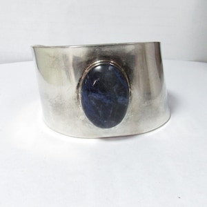 May include: A wide silver cuff bracelet featuring a large, polished, oval-shaped dark blue sodalite gemstone set in the center. The metal surface has a smooth, reflective finish with subtle variations in tone across the curved band.