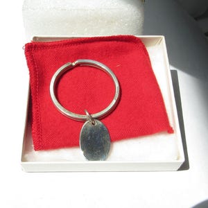 May include: A silver-toned metal keychain featuring a circular split ring and a smooth, oval-shaped pendant. The keychain is presented inside a white box containing a bright red fabric square, providing a clear contrast to the metallic finish.