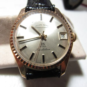 May include: A vintage gold-toned watch with a silver face, black leather band, and a date display. The watch face features the text "FELIPE RENE SUPERAUTOMATIC", "25 RUBIS Revelation", and "SWISS MADE". The watch has a fluted bezel.