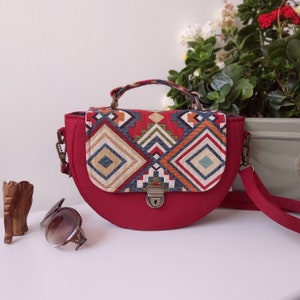 May include: A red half-moon shaped handbag with a geometric patterned flap and a top handle. The bag has a metal clasp and a long adjustable strap.