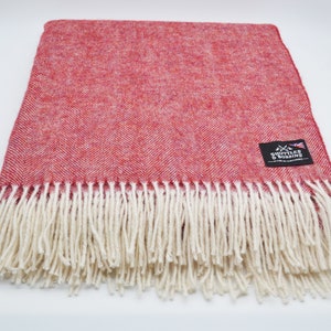 Op de afbeelding: Een rode herringbone deken met een witte franje. De deken heeft een geweven label met de tekst "Shottle & Borrins" met een kleine Britse vlag.
