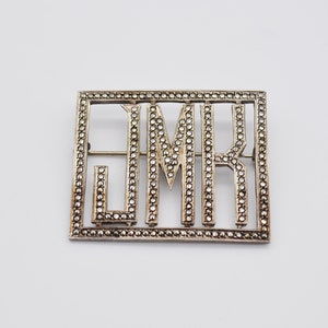 May include: A rectangular silver-toned brooch featuring the letters JMR. The letters are outlined and filled with small, sparkling stones. The brooch has a pin closure on the back and is set against a white background.