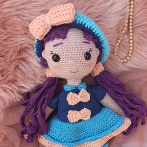 Puede incluir: Una muñeca de crochet con pelo morado, un sombrero azul y melocotón, un vestido azul con lazos de melocotón y medias a rayas azules y blancas. La muñeca lleva zapatos azules con lazos de melocotón.
